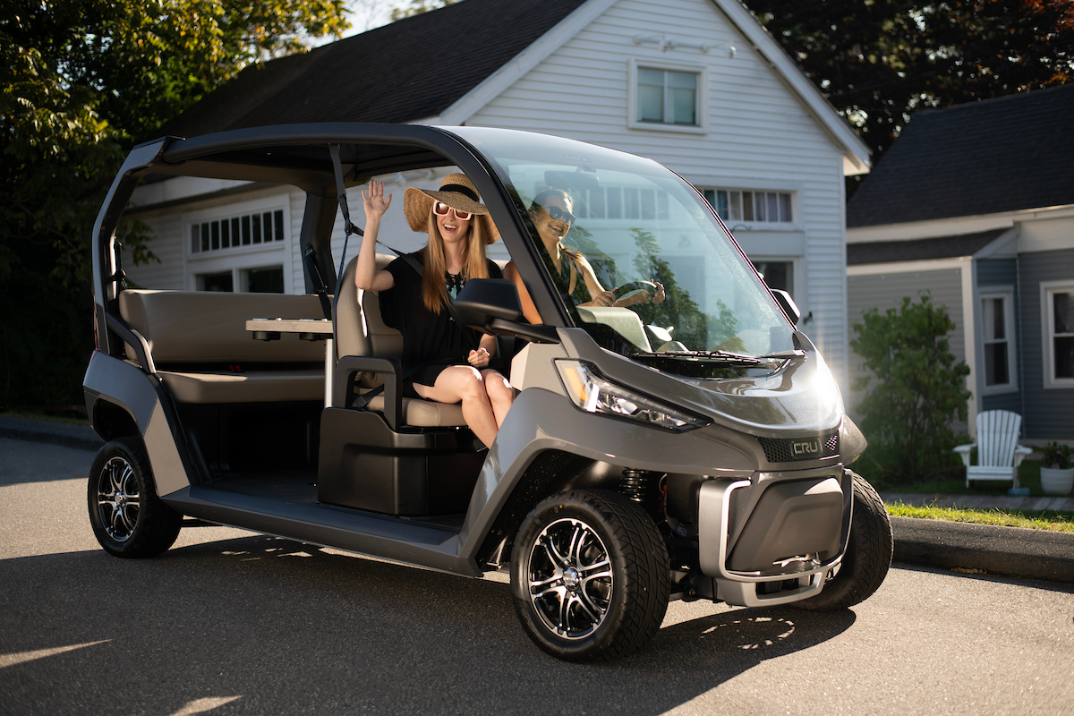 Club Car Cru purchased from an authorized Club Car Dealer with two passengers driving on a residential street on a sunny day.